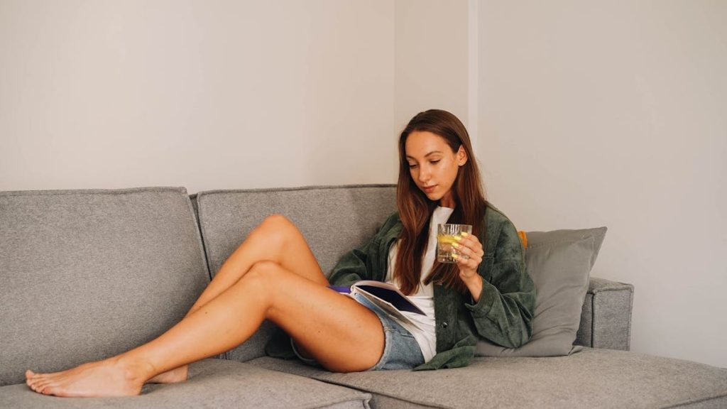 Woman reading a book on a grey sofa while holding a glass of water in a relaxed indoor setting.